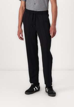 Pantalones negros, ligeros y con cintura elástica, de corte relajado. Presentan discretas rayas verticales y un detalle de cordón. Combinados con zapatillas negras.