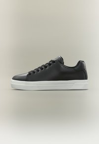 Μαύρο δερμάτινο αθλητικό παπούτσι χαμηλού ύψους με μαύρα κορδόνια και παχιά λευκή λαστιχένια σόλα, εμφανιζόμενο σε προφίλ σε ουδέτερο φόντο.