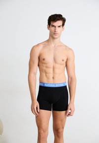 Μαύρα μποξέρ σλιπ με μπλε ελαστική ζώνη που φέρει το κείμενο "BJÖRN BORG". Λείος ύφασμα με εφαρμοστό σχέδιο και εκτεθειμένα άκρα ποδιών.