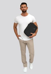 Custodia nera per laptop con una finitura liscia, portata da un uomo in una t-shirt bianca e pantaloni beige, in piedi contro uno sfondo grigio.