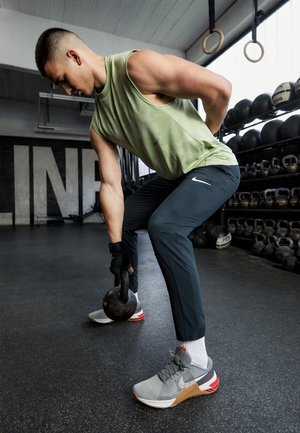 Homme soulevant une kettlebell dans une salle de sport avec des étagères de poids et des anneaux de gymnastique en arrière-plan.