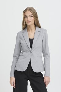 Jonge vrouw met lang blond haar, gekleed in een lichtgrijze getailleerde blazer over een zwarte top en zwarte broek, tegen een effen witte achtergrond.