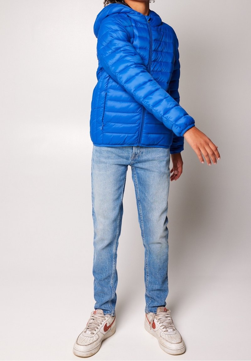 Chaqueta de plumas azul con capucha, con un diseño acolchado horizontal y cierre de cremallera. Combinada con jeans azul claro y zapatillas blancas.