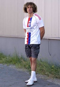 Maillot de soccer blanc avec des rayures verticales bleues et rouges, shorts cargo gris, chaussettes blanches et chaussures blanches. Cheveux bouclés, debout à l'extérieur.