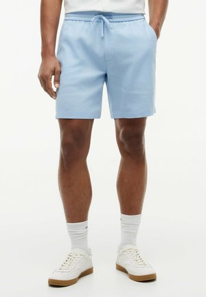 Mężczyzna ubrany w jasnoniebieskie szorty ze sznurkiem, białe skarpetki do kostek oraz białe tenisówki, stojący z rękami w kieszeniach na białym tle.