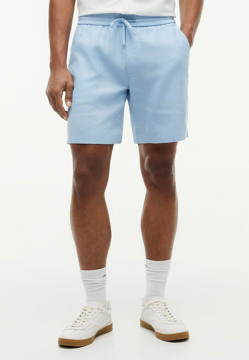 Mężczyzna ubrany w jasnoniebieskie szorty ze sznurkiem, białe skarpetki do kostek oraz białe tenisówki, stojący z rękami w kieszeniach na białym tle.