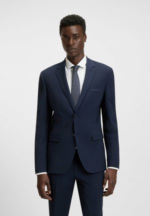 Ein Mann mit kurzen, lockigen Haaren steht in einem gut sitzenden marineblauen Anzug und Krawatte und zeigt einen selbstbewussten Gesichtsausdruck vor einem schlichten weißen Hintergrund.