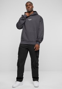 Grå hettegenser med lomme foran og hvit logo, sammen med svarte cargo-bukser med flere lommer og hvite sneakers.