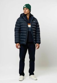 Marineblauwe gewatteerde jas met capuchon, rits aan de voorkant en een gequiltde textuur. Draag het over een veelkleurige gestreepte trui en gecombineerd met donkere jeans.