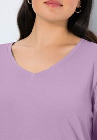 Žena s tmavými vlasy, oblečená v stříbrných kruhových náušnicích a levandulovém tričku s výstřihem do V, zachycena od krku až k horní části hrudi na bílém pozadí.