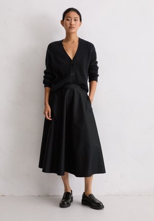 Zwarte gebreide cardigan met een V-hals en knoopsluiting, gecombineerd met een zwarte rok met hoge taille en een vloeiende pasvorm, inclusief zakken. Zwarte schoenen.