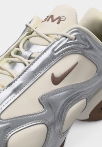 Αθλητικό παπούτσι με μπεζ και ασημένιες επιφάνειες, καφέ λογότυπο Nike, μπεζ κορδόνια και κείμενο "Zoom" στη γλώσσα, εμφανιζόμενο από κοντινή γωνία.