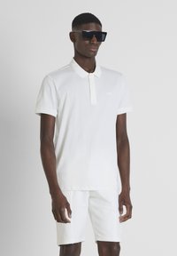 Polo blanco de algodón con cuello, que presenta un pequeño logotipo bordado. Combinado con pantalones cortos blancos a juego. Modelo luciendo gafas de sol oscuras.