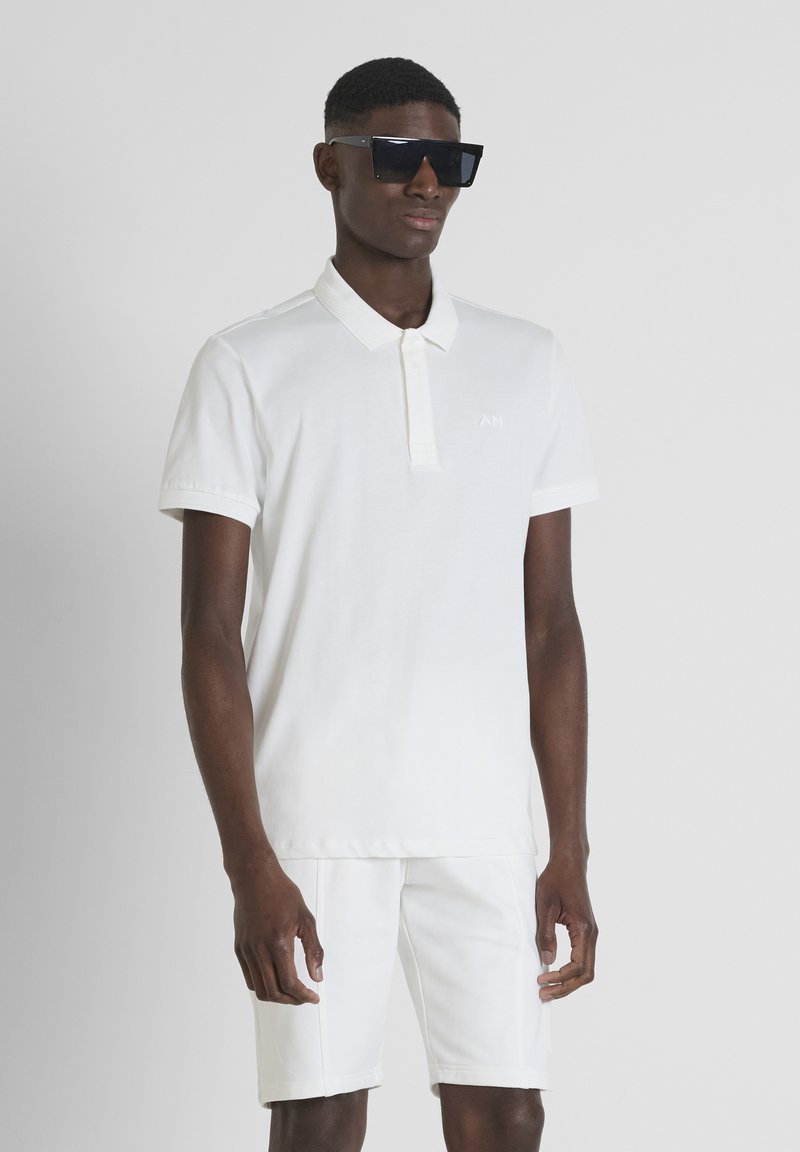 Polo blanco de algodón con cuello, que presenta un pequeño logotipo bordado. Combinado con pantalones cortos blancos a juego. Modelo luciendo gafas de sol oscuras.