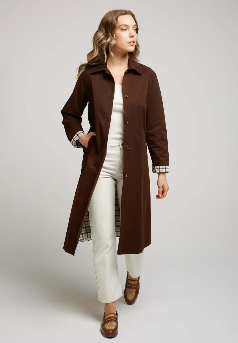 Manteau long marron avec un col et une fermeture par boutons, présentant une doublure à motif à carreaux sur les poignets. Porté par-dessus un haut blanc et un pantalon crème.