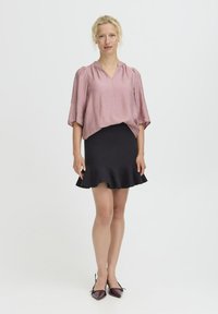 Hellrosa Bluse mit kurzen Ärmeln, V-Ausschnitt und gerafftem Detail, kombiniert mit einem schwarzen, ausgestellten Minirock und kastanienbraunen, spitzen Ballerinas.