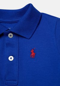 Modrá polo košile vyrobená z bavlny, s žebrovaným límcem a zapínáním na dva knoflíky. Vyšívané červené logo na levé straně hrudi.