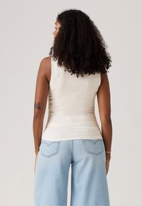 Frau mit lockigem Haar, die ein ärmelloses beigefarbenes Oberteil und leichte weite Jeans in Hellblau trägt, stehend mit dem Rücken zu einer schlichten Hintergrund.