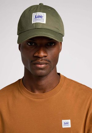 Olivgrüner Baseball-Cap mit einem quadratischen weißen Patch, auf dem das blaue "Lee"-Logo abgebildet ist. Getragen mit einem braunen T-Shirt, in einem Nahaufnahme-Porträtstil.