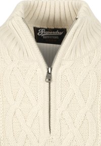 Kremowy sweter w splot warkoczowy z wysokim kołnierzem, z srebrnym zamkiem błyskawicznym i teksturowanym wzorem, oznaczony napisem "Superdry Outfitters".