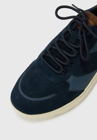 Navy blue suede sneaker with a textured finish, featuring contrasting lighter accents, round laces, and a white rubber sole.