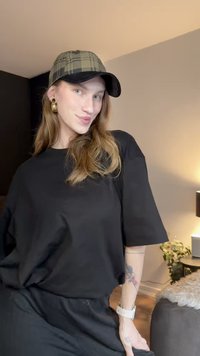 Fekete túlméretezett póló, sima anyagból, bő szabással, zöld kockás sapkával párosítva. Figyelemre méltó arany fülbevalók és fehér karóra a csuklón.