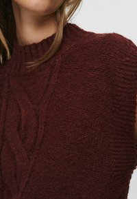 Artimiausia nuotrauka rodo asmenį, vilkintį tekstūrinį, tamsiai bordo spalvos megztinį su aukštu, raštuotu kaklu.