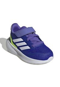 Fialová sportovní bota se síťovinovou svrchní částí, bílými akcenty a suchým zipem. Má strukturovanou podrážku a jasně zelenou boční panel.