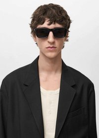 Occhiali da sole neri con lenti scure, indossati da una persona in un blazer nero sopra una maglietta chiara con bottoni, che promuove uno stile minimalista.