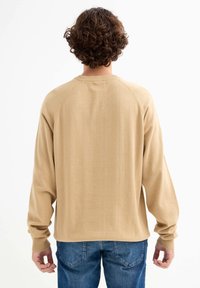 Suéter de punto beige con mangas raglán, puños y cuello acanalados, textura suave, llevado sobre unos jeans de mezclilla azul, visto desde la parte posterior.
