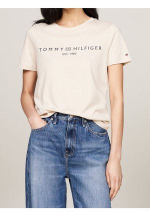 Beżowa bawełniana koszulka z okrągłym dekoltem, z nadrukiem "TOMMY HILFIGER EST. 1985" w granatowym kolorze, zestawiona z wysokim stanem niebieskich dżinsów.