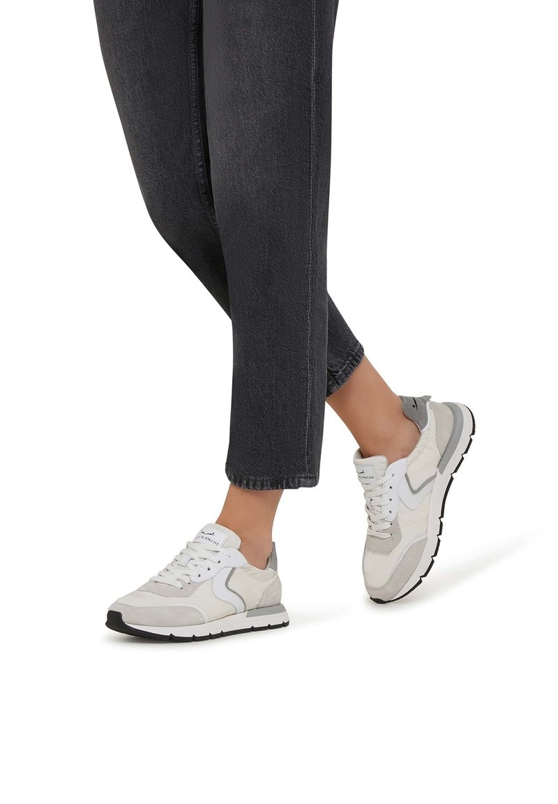 Jambes portant un pantalon noir court et des baskets blanc-gris avec semelles noires, montrées en mouvement de marche sur fond blanc.