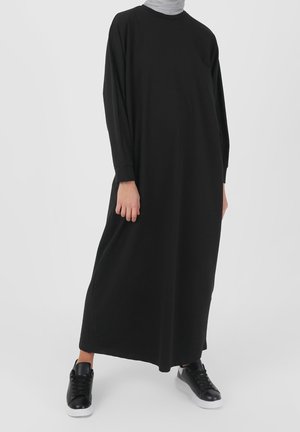 Person trägt ein langes schwarzes Kleid mit langen Ärmeln, kombiniert mit schwarzen Turnschuhen mit weißen Sohlen und steht vor einem einfarbigen Hintergrund.