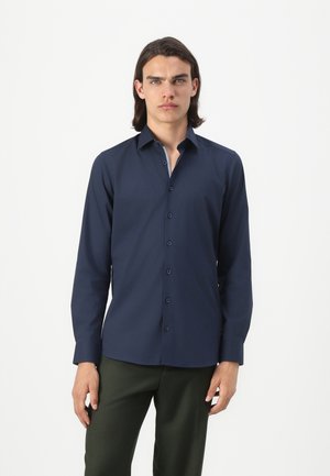 Chemise à manches longues bleu marine avec col boutonné, présentant une texture lisse et une coupe standard, associée à un pantalon vert foncé.