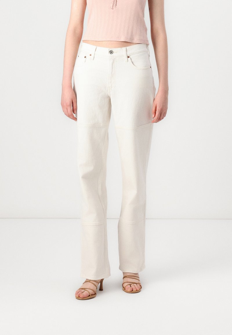 White denim pants with a straight-leg design, featuring seams and stitched detailing. Paired with a light pink ribbed top and strappy sandals.