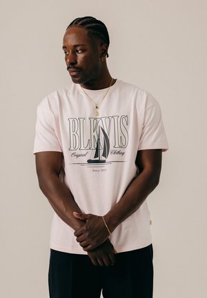Homme aux cheveux tressés portant un t-shirt rose BLK IN IS avec un motif de voilier, des colliers en or et un bracelet, regardant vers la gauche sur un fond uni.