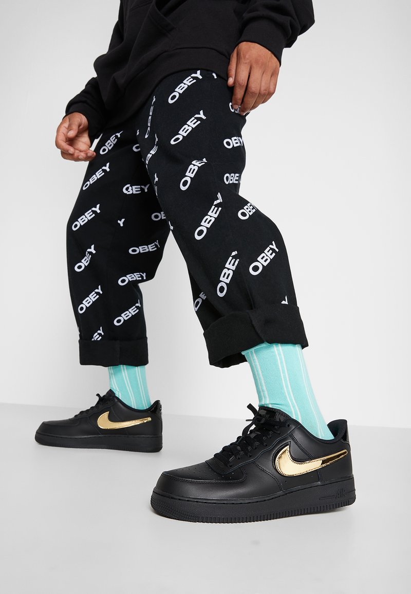 Personne portant un pantalon noir avec des logos blancs "OBEY", des chaussettes rayées turquoise et des baskets noires avec un logo Nike doré sur un sol gris.