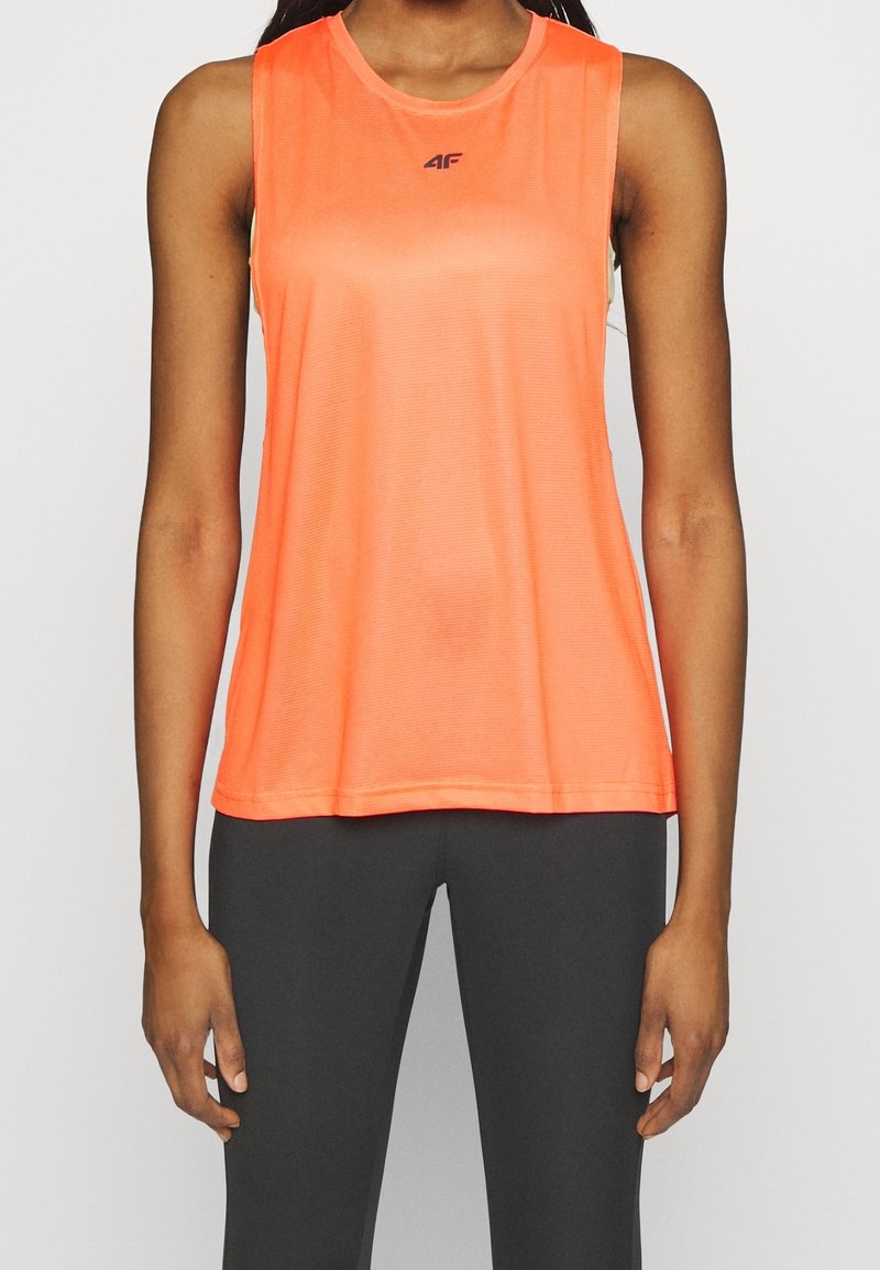 Femme portant un débardeur de sport orange vif sans manches et un legging foncé ajusté, debout devant un fond clair uni.