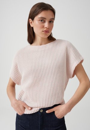 Giovane donna con capelli castani lisci, indossa una maglietta rosa a maniche corte lavorata a maglia con trama e jeans blu scuro con le mani in tasca.