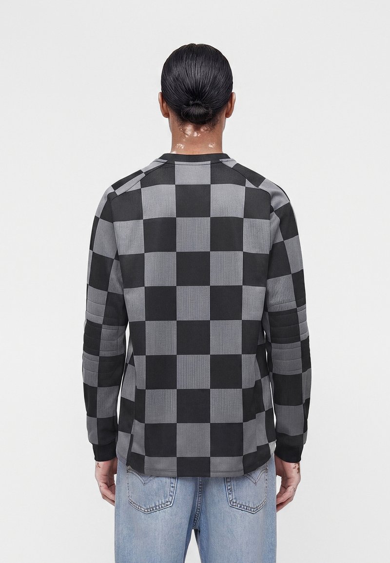 Person mit zurückgebundenen Haaren, die ein schwarz-grau kariertes Langarmhemd und hellblaue Jeans trägt, von hinten vor einem einfarbigen Hintergrund gesehen.