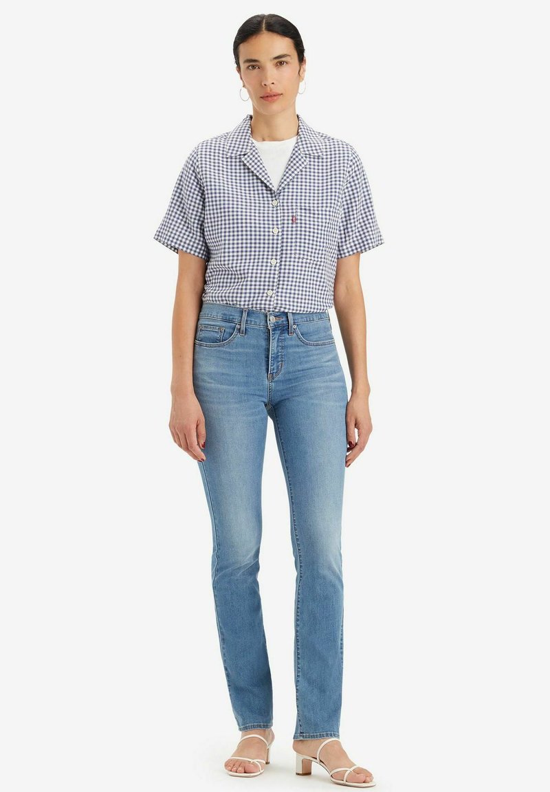 Camicia a maniche corte a quadri blu e bianca, abbinata a jeans azzurri chiari e sandali bianchi con cinturino. Il modello è posizionato davanti a uno sfondo neutro.