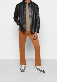 Czarna skórzana kurtka z zatrzaskami, pod spodem beżowa koszula w paski, brązowe spodnie oraz wielokolorowe buty sportowe z niebieskimi i żółtymi akcentami.