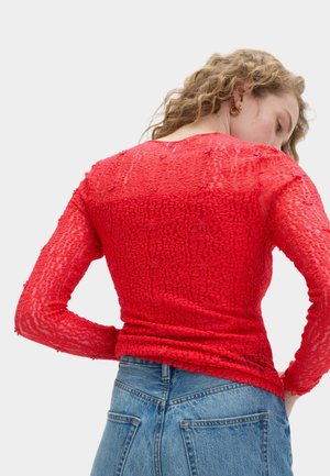 Top a maniche lunghe in pizzo rosso con finitura testurizzata, caratterizzato da una silhouette aderente. Abbinato a jeans di denim blu a vita alta.