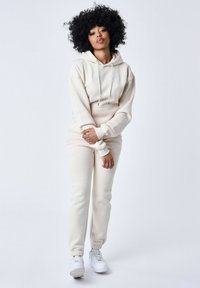 Sudadera con capucha beige y joggers a juego, hechos de una tela suave, que cuentan con cordón, puños acanalados y un logo bordado. Las zapatillas completan el look.