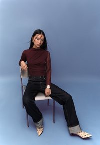 Viininpunainen pitkähihainen paita, jossa on kerätty yksityiskohta, yhdistettynä väljään tummiin farkkuihin ja kuvioituihin, teräväkärkisiin korkokenkiin, istuen metallisella tuolilla.