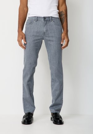 Mann trägt hellgraue Jeans mit geradem Bein, schwarze Loafers und ein weißes ärmelloses Shirt, steht vor einem einfachen hellen Hintergrund.