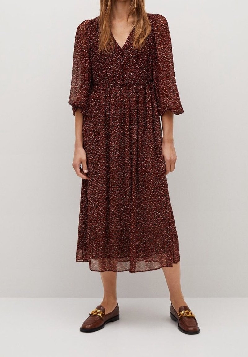 Femme portant une robe midi fleurie bordeaux avec des manches bouffantes transparentes trois-quarts et des mocassins marron avec un détail chaîne dorée.