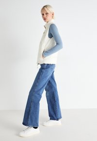 White puffy vest with a high collar, worn over a light blue long-sleeve top. Paired with wide-leg blue jeans and white sneakers.