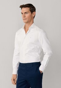 Mann mit braunen Haaren, der ein weißes Hemd mit Knöpfen und dunkelblaue Hosen trägt, steht mit einer Hand in der Tasche vor einem neutralen Hintergrund.