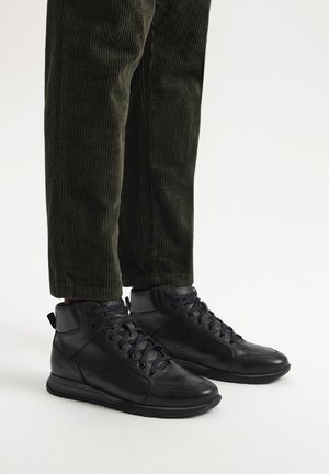 Czarne skórzane sneakersy za kostkę z płaskimi sznurowadłami, teksturowaną cholewką, wyściółką w kołnierzu i gumową podeszwą, w połączeniu z ciemnozielonymi spodniami z korduroju.